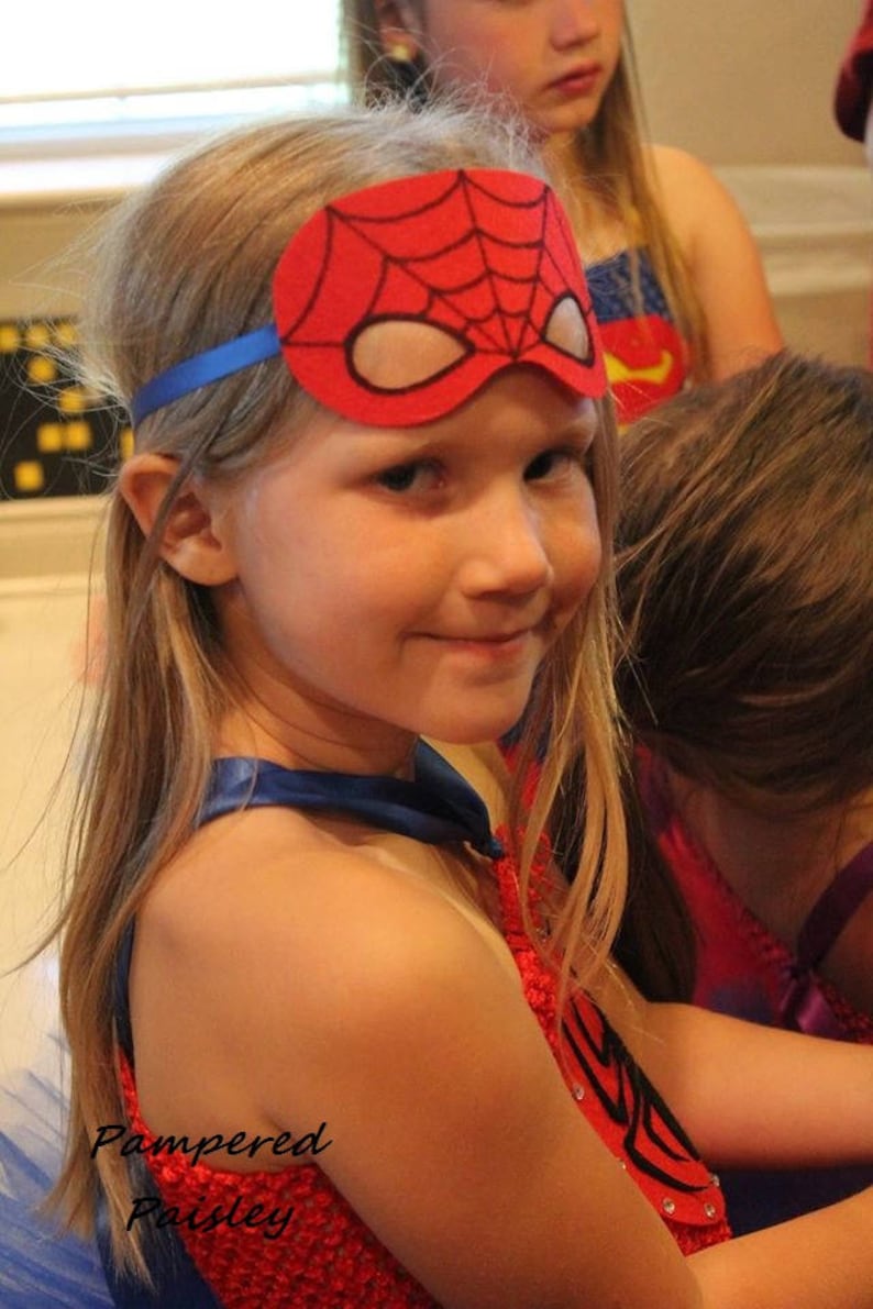 May include: A red felt superhero mask featuring a black spiderweb pattern and white eye cutouts, secured with a blue elastic strap. The mask is worn across the forehead, paired with a red textured bodice and a blue cape with a blue neck tie.