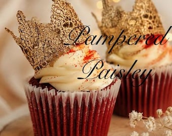 May include: Two red velvet cupcakes in white paper liners, each topped with a swirl of white frosting, a dusting of red powder, and a decorative gold lace crown. The text Pampered Paisley is overlaid in black script across the center of the image.