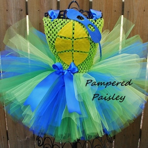 May include: A green and blue tutu dress with a matching eye mask. The bodice is crocheted in green with a yellow turtle shell design. Blue satin ribbons and layers of tulle create the skirt. The text "Pampered Paisley" is visible.