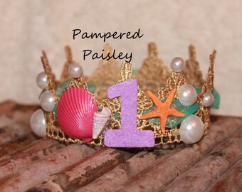 May include: A gold lace crown featuring the text Pampered Paisley, a glittery purple number one, a bright pink seashell, a small white spiral shell, an orange starfish, and several white pearl-like beads attached to the metallic gold trim.