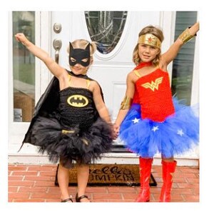 May include: A black Batman costume featuring a mask, cape, and tutu skirt with a yellow logo, alongside a red and blue Wonder Woman costume with a gold headband, arm cuffs, and a blue tutu skirt adorned with white stars, both worn as superhero outfits.