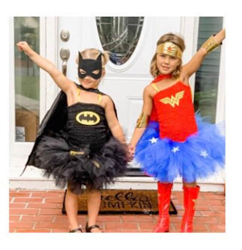 May include: A black Batman costume featuring a mask, cape, and tutu skirt with a yellow logo, alongside a red and blue Wonder Woman costume with a gold headband, arm cuffs, and a blue tutu skirt adorned with white stars, both worn as superhero outfits.