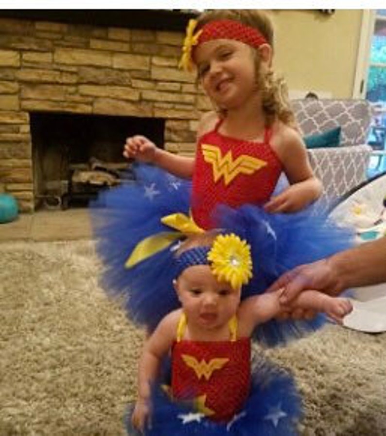 May include: Two matching Wonder Woman themed outfits consisting of red crochet tops with yellow W logos, blue tulle skirts with white star accents, and coordinating headbands featuring yellow flowers, designed for two different sized individuals.