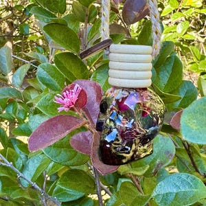 May include: A skull-shaped glass car air freshener bottle filled with dried pink and purple flower petals, topped with a ribbed light-colored wooden cap and suspended by a braided cord with metallic gold threading.