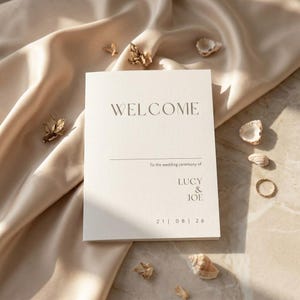 May include: A rectangular white wedding program card with black text that reads WELCOME, To the wedding ceremony of, LUCY & JOE, and the date 21 | 08 | 26. The card is centered on a smooth beige fabric with small seashells and a gold ring.