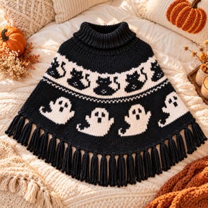 Puede incluir: Un poncho de punto negro con cuello alto y dobladillo con flecos. La prenda presenta un patrón repetitivo de fantasmas blancos en la parte inferior y una banda de gatos negros sobre fondo blanco en la zona del pecho.