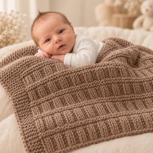 May include: A baby with light skin and brown hair wearing a white long-sleeved ribbed shirt, resting on a thick, hand-knitted brown blanket. The blanket features a textured pattern of alternating garter stitch borders and vertical ribbed columns.