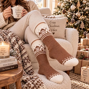 May include: A pair of brown and cream knit socks featuring a repeating pattern of small houses around the cuffs and white toe sections. The socks are made of a thick, textured yarn and reach mid-calf, displaying a cosy, winter-inspired design.