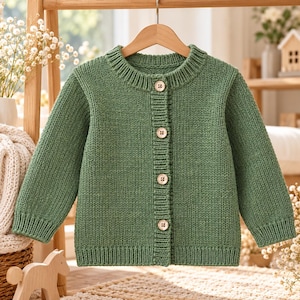 Può includere: Un cardigan in maglia a maniche lunghe color verde salvia con girocollo a coste, polsini a coste e orlo a coste. Il capo si chiude sul davanti con quattro bottoni in legno di colore chiaro ed è esposto su una gruccia di legno.