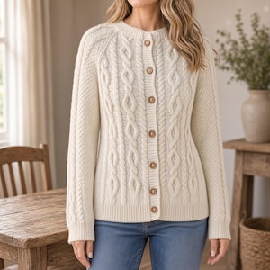 May include: A cream-coloured cable knit cardigan featuring a crew neckline, long sleeves, and a front button closure with six round brown buttons. The garment displays a textured pattern of vertical cable twists and side panels with a honeycomb stitch.