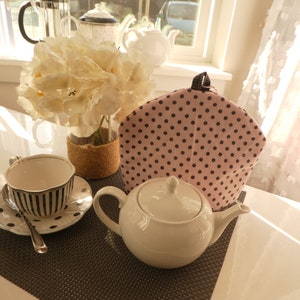 May include: A white teapot with a pink and black polka dot tea cozy on a black and white placemat. A white teacup with black stripes is on a black and white saucer with a pink polka dot design.