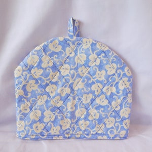 May include: A blue and white quilted tea cozy with a floral pattern. The cozy has a loop handle on top.