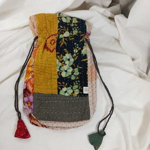 Può includere: Una borsa a cordoncino patchwork con una varietà di motivi floreali in tonalità di giallo, verde, blu e rosa. La borsa ha un fondo in tessuto grigio e un piccolo cartellino con la lettera "S" su di esso.