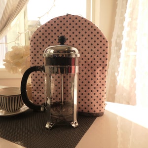 May include: A silver French press coffee maker with a black handle and a pink and black polka dot fabric cover. The coffee maker is sitting on a brown woven placemat.