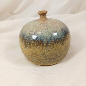 May include: A handcrafted ceramic vase with a rounded body and a narrow neck. The vase features a blend of colors, including tan, blue, and brown, with a glossy finish. The vase is a decorative item.