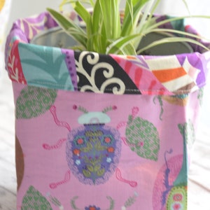 May include: A pink fabric planter with a colourful patterned trim and a design of stylised bugs. The planter is filled with a green plant.
