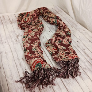 May include: A long, brown paisley patterned scarf with fringed ends. The scarf is made of a soft, lightweight fabric and features a paisley design in shades of red, green, and cream.