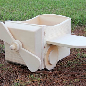 Puede incluir: Un avión de juguete de madera con una hélice, un pequeño compartimento de almacenamiento y una rueda. El juguete está hecho de madera natural y tiene un color marrón claro.