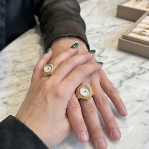 May include: Two gold-toned ring watches featuring round white dials with black numerals and fluted bezels. Each ring has a metallic band and a small winding crown on the side, designed to be worn on the finger as a functional timepiece.