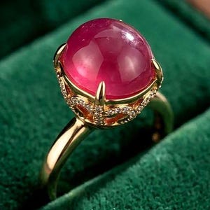 May include: A gold ring featuring a large, smooth, cabochon-cut deep pink gemstone set in a decorative basket setting. The setting is adorned with small, clear, sparkling stones along the gallery, supported by a polished gold band.