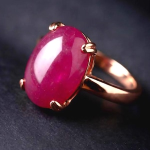 May include: A ring featuring a large, oval-shaped, cabochon-cut magenta gemstone set in a polished rose gold band. The stone is secured by four prongs, showcasing a smooth, rounded surface with a reflective sheen and deep pinkish-red colour.