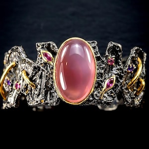 Puede incluir: Un brazalete con una gran piedra cabujón ovalada de color rosa translúcido engastada en un bisel dorado. La banda tiene un acabado metálico oscuro, oxidado y texturizado con detalles de alambre dorado y pequeñas piedras facetadas incrustadas.