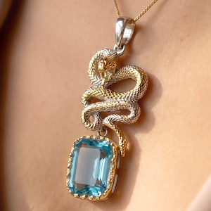 Peut inclure: Un pendentif collier représentant un serpent enroulé avec une finition bicolore or et argent, placé au-dessus d'une grande pierre précieuse rectangulaire bleu clair à facettes, sertie dans un cadre doré, suspendu à une fine chaîne en or.