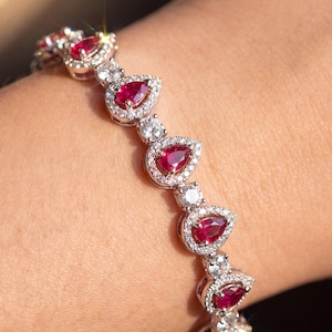 May include: A silver-toned bracelet featuring a series of pear-shaped red gemstones, each surrounded by a halo of small clear stones. The links between the pear-shaped settings are accented with single round clear stones, creating a continuous design.