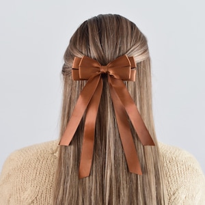 May include: A brown satin hair bow with long, flowing ribbons is secured in the hair. The bow is a rich, warm brown color and adds a touch of elegance. The hair is long and light brown, with a sweater in the background.