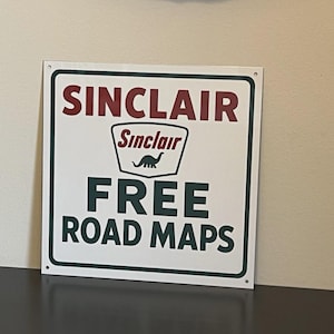 May include: A square white metal sign with a dark green border and four corner mounting holes. The sign features the red text SINCLAIR at the top, a central Sinclair logo with a dinosaur graphic, and the dark green text FREE ROAD MAPS below.