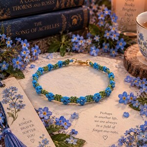 May include: A handmade bracelet featuring a repeating pattern of small, blue, flower-shaped knots interspersed with green cord segments. The bracelet is secured with a gold-toned metal lobster clasp and is displayed on a surface surrounded by blue flowers.
