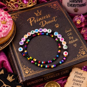 May include: A beaded necklace featuring white round beads with colorful letters spelling out GODDAMMIT DONUT, interspersed with small black beads and a variety of multicolored accent beads arranged in a circular pattern on a dark book cover.