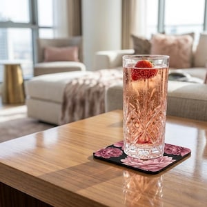 Könnte beinhalten: Ein hohes, klares, facettiertes Glas gefüllt mit einem rosa kohlensäurehaltigen Getränk und einer frischen Erdbeere, das auf einem quadratischen Untersetzer mit dunklem Hintergrund und rosa-burgunderfarbenem Blumenmuster steht.
