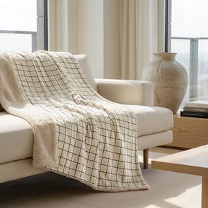 Könnte beinhalten: Eine cremefarbene Wohndecke mit schwarzem Karomuster auf der einen Seite und einem strukturierten, fleeceartigen Material auf der Rückseite. Die Decke liegt über einem hellen Sofa und zeigt ihre weiche, flauschige Textur und das geometrische Design.
