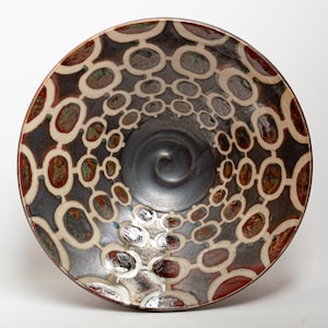 May include: A round ceramic bowl featuring a dark gray center with a spiral pattern. The interior walls are decorated with a repeating cream-colored chain-link design over a dark background, with accents of metallic copper and green glaze.