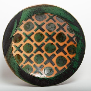 May include: A round ceramic plate featuring a central grid pattern of dark brown X shapes and orange squares containing dark green circular spots. The outer rim of the plate is a wide band of mottled dark green and black glaze.