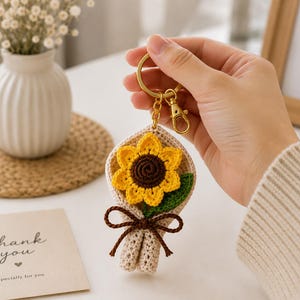 以下が含まれることがあります： 黄色と茶色の花びらに緑の葉がついた、かぎ針編みのひまわりのキーホルダー。ベージュのかぎ針編みのブーケスリーブに包まれ、ダークブラウンの毛糸の結び目がついており、ゴールドトーンの金属製リングとロブスタークラスプに取り付けられています。