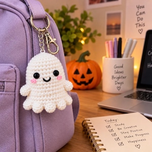 May include: A white crocheted ghost keychain with black plastic eyes, a stitched black mouth, and pink embroidered cheeks. The ghost is attached to a silver-toned metal keyring and lobster clasp, hanging from the side of a light purple backpack.
