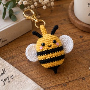 Puede incluir: Un llavero de abeja tejido a ganchillo con cuerpo de rayas amarillas y negras, dos alas blancas, antenas negras y ojos de cuentas negras con mejillas rosadas bordadas. La abeja está unida a un mosquetón de metal dorado y una argolla.