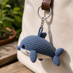 Peut inclure: Un porte-clés dauphin en crochet bleu et blanc avec une petite nageoire dorsale, des nageoires latérales et une queue. Le dauphin possède un œil en perle noire et un sourire brodé. Il est fixé à un anneau en métal argenté par une chaînette.