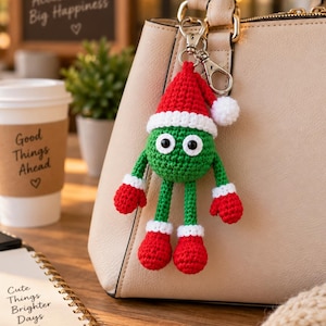 Op de afbeelding: Een gehaakte sleutelhanger met een rond groen figuurtje met een rode kerstmuts, witte rand, rode wanten en rode laarsjes. Het figuurtje heeft twee zwarte ogen en is bevestigd aan een zilverkleurige metalen karabijnhaak aan een beige tas.