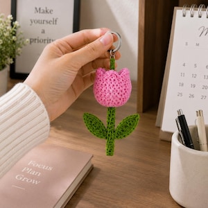 May include: A crochet tulip keyring featuring a bright pink flower head with a textured green stem and two green leaves attached to a silver metal ring. The handcrafted floral design is made from yarn and displays a detailed stitch pattern.
