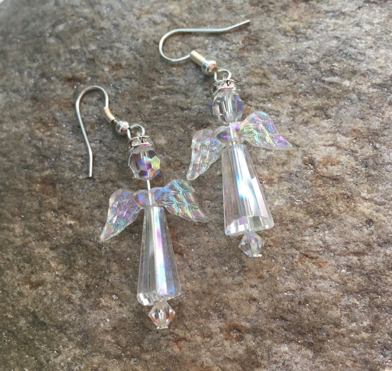 May include: A pair of silver dangle earrings featuring crystal angel charms. The angels have iridescent wings and clear bodies.