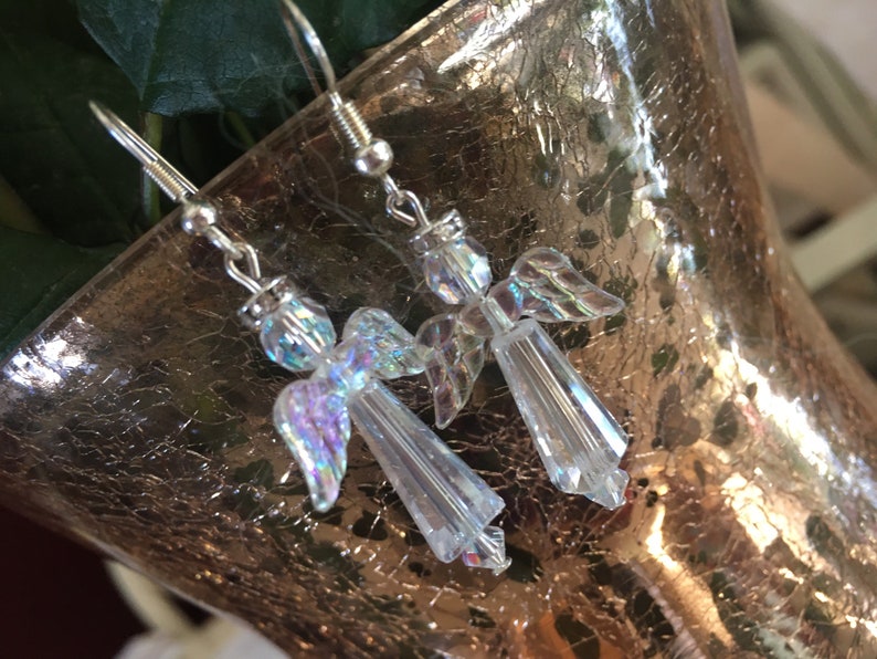 May include: A pair of silver dangle earrings featuring iridescent angel charms. The angels are made of clear crystal with iridescent wings.