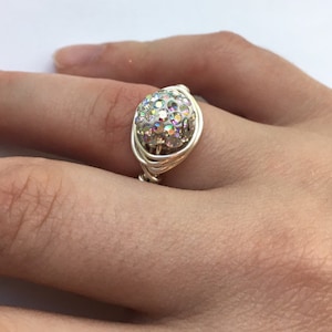 May include: A silver wire-wrapped ring featuring a large, iridescent crystal ball. The crystal ball is composed of numerous smaller, colorful crystals. The ring is shown on a person's finger.