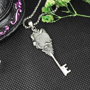 May include: A silver-toned metal pendant shaped like a key, featuring a circular head with a labyrinthine spiral pattern surrounded by two coiled snakes. The pendant hangs from a silver-toned ball chain necklace.