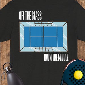 May include: A black t-shirt featuring a graphic of a blue padel court viewed from above, with the text "OFF THE GLASS" above the court and "OWN THE MIDDLE" below it. A black water bottle and a black padel racket with a yellow ball are visible.