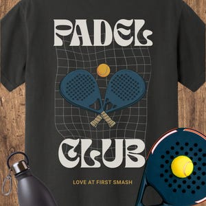 May include: A black t-shirt featuring a graphic with the text PADEL CLUB and LOVE AT FIRST SMASH. The design includes two crossed dark blue padel rackets and a yellow ball over a grid pattern. A separate dark blue padel racket and water bottle.