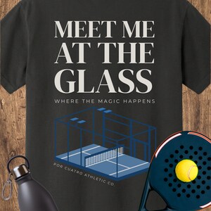 May include: A black t-shirt featuring the white text MEET ME AT THE GLASS WHERE THE MAGIC HAPPENS above a blue graphic of a padel court and the text POR CUATRO ATHLETIC CO. A black water bottle and a black padel racket with a yellow ball are present.