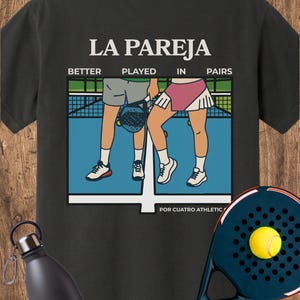 May include: A black t-shirt featuring a graphic of two people on a blue padel court with the text LA PAREJA, BETTER PLAYED IN PAIRS, and POR CUATRO ATHLETIC. A dark blue padel racket with a yellow ball and a black water bottle are also visible.